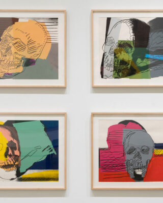 Four framed artworks of colorful skulls are displayed in two rows on a white wall. Each piece combines vibrant hues and abstract shapes.